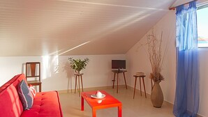 Una sala de estar acogedora, con un sofá rojo, una pequeña mesa con un objeto blanco, un televisor sobre un mueble bajo y un jarrón grande con plantas.