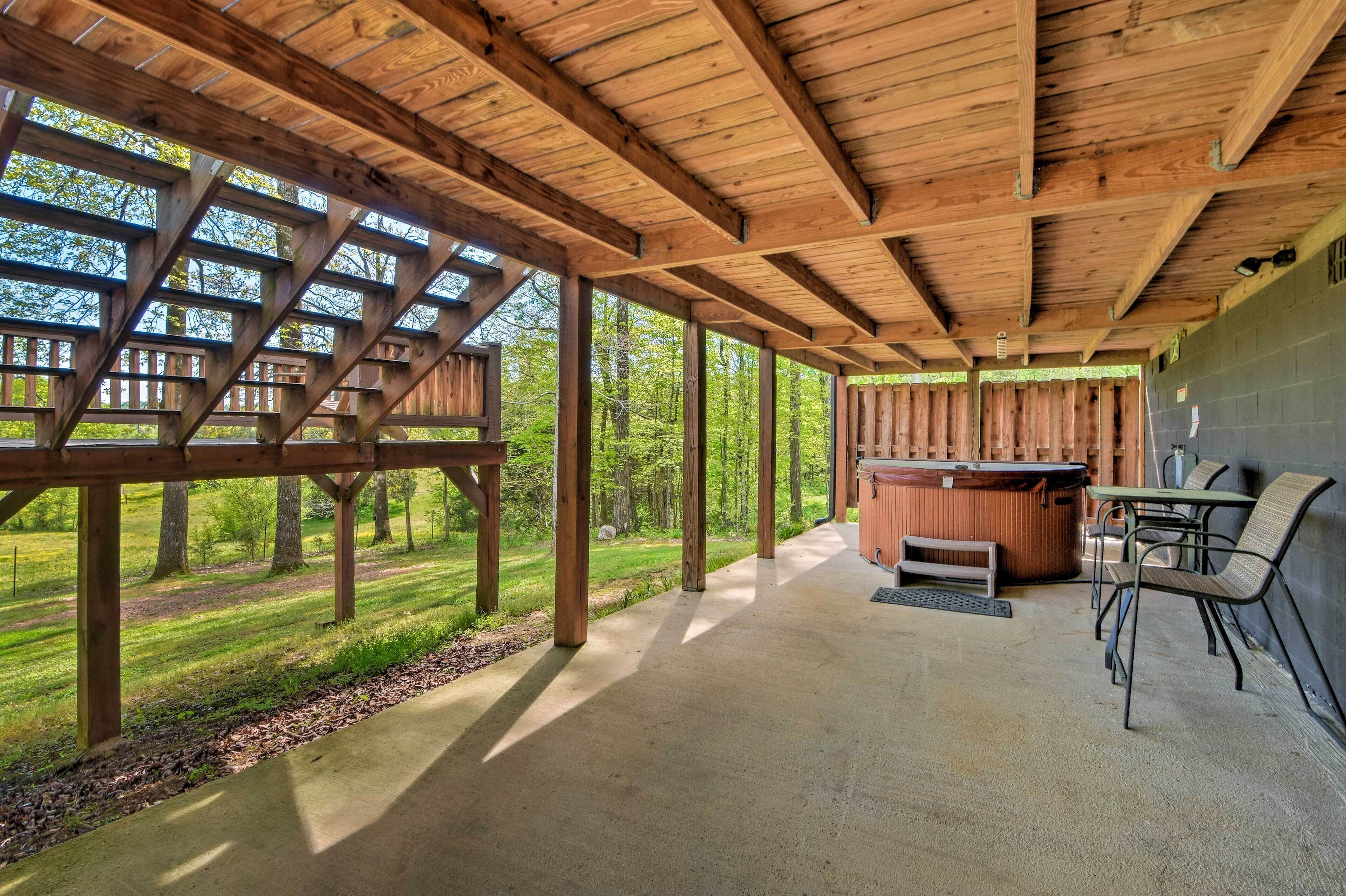 A covered outdoor area with a hot tub, seating, and a view of a forested area.