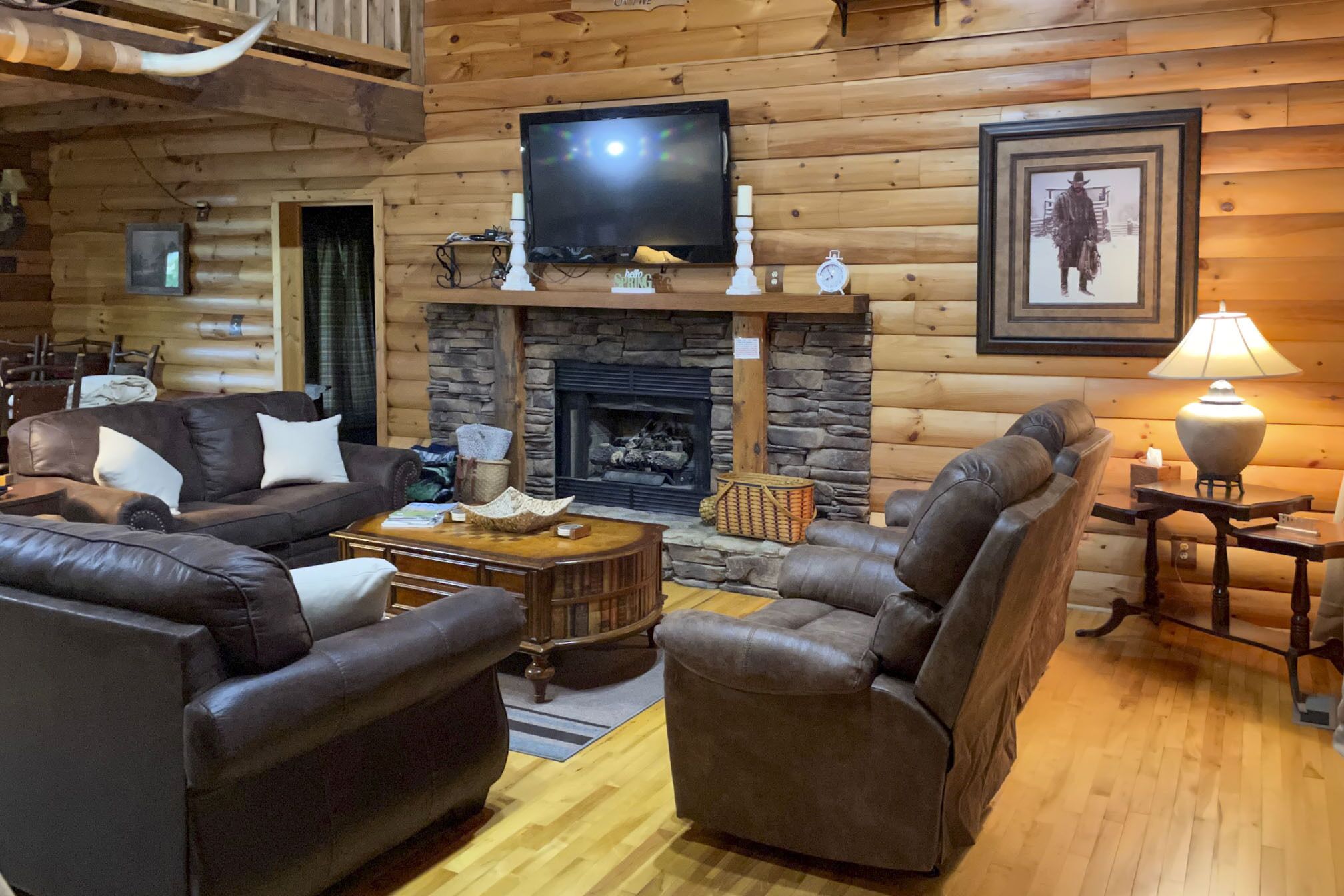 A cozy log cabin interior with a fireplace, leather furniture, and a television mounted above it.