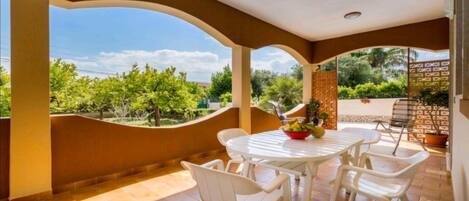 Un espace extérieur aménagé, abrité, avec une table et des chaises, un saladier rempli de fruits et une vue sur la verdure et les arbres.