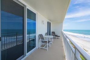 A balcony with a view of the beach, a table with chairs, and a small table with a chair.