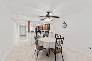 A dining area with a round table, four chairs, a ceiling fan, and a kitchen in the background.