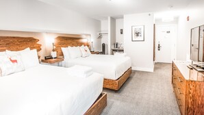 A hotel room with two beds, a wooden headboard, a desk with a chair, a TV, and a bathroom door. - Basecamp Lodge I Double Queen Room (Canmore)