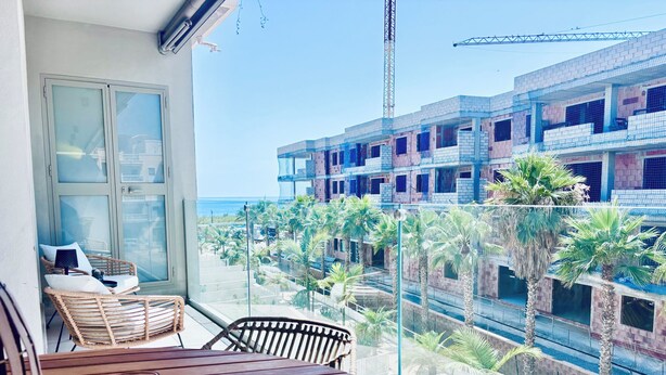 A balcony with wicker furniture, a glass table, and a view of a construction site with palm trees. - Stylish apartment with sea views (Torrox)