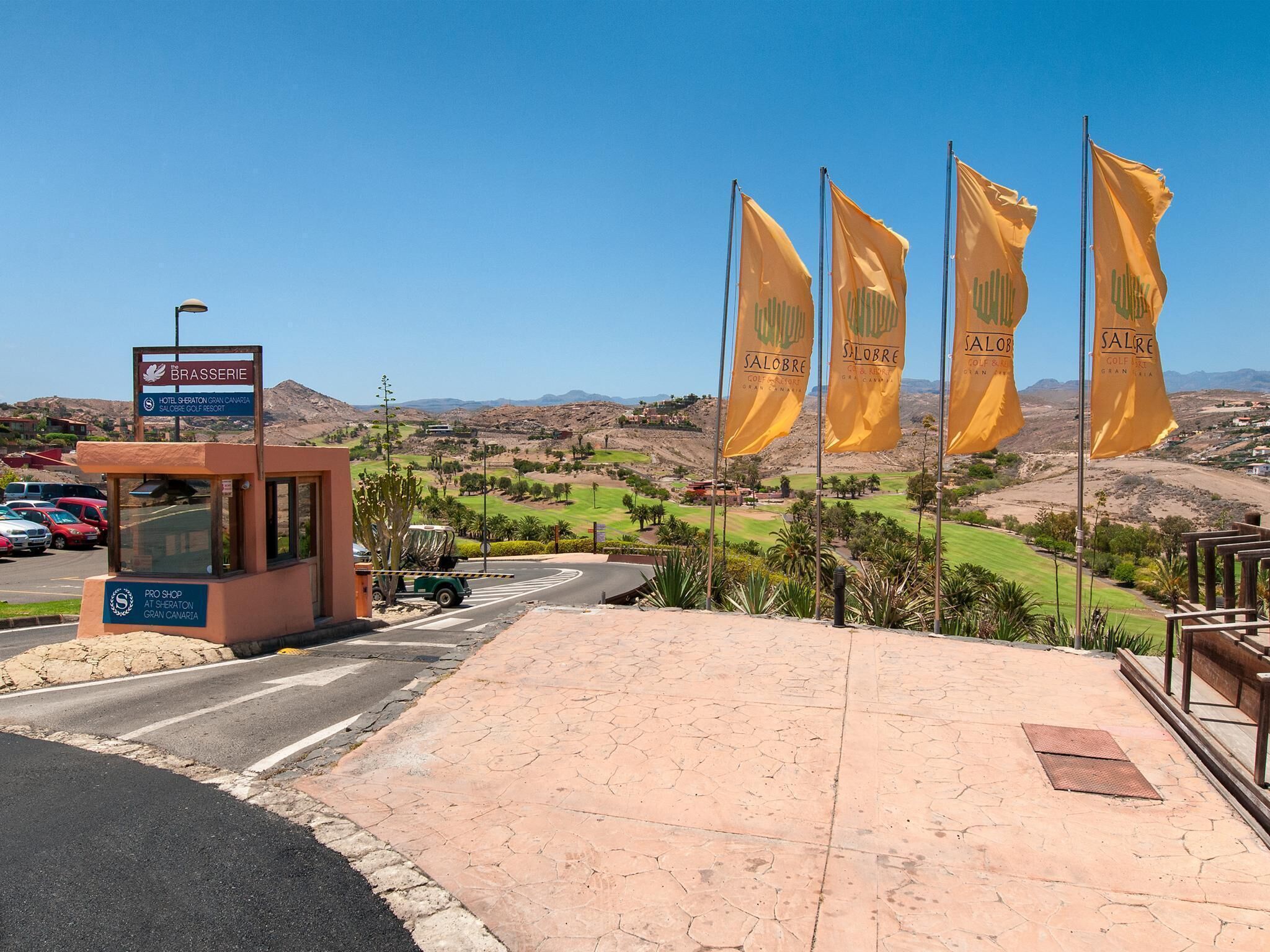 Entrada de hotel con un letrero que indica Brasserie, un campo de golf al fondo y varias banderas amarillas.