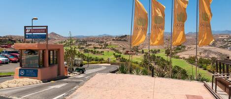 Entrada de hotel con un letrero que indica Brasserie, un campo de golf al fondo y varias banderas amarillas.