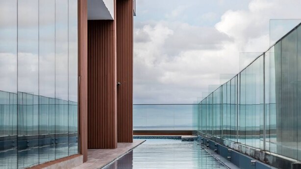 Ein moderner Infinity-Pool mit Glasgeländern, einer Holzsäule und Blick aufs Meer.