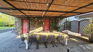 An outdoor covered patio with a stone wall, a checkered table, and wicker chairs. - Refined and welcoming Villa with private pool surrounded by nature (Omignano Scalo)