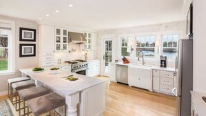 A modern kitchen with white cabinetry, a central island with stools, stainless steel appliances, and a view of a lake through large windows. - Cape Cod Intellectual Property (Eastham)