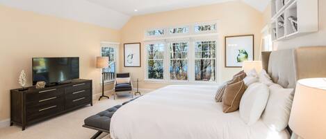 A spacious bedroom with a large bed, a TV, a dresser, and a window with a view of trees.