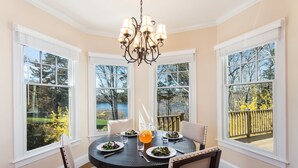 A dining room with a round table, chairs, a chandelier, and large windows with a view of trees and a deck. - Cape Cod Intellectual Property (Eastham)