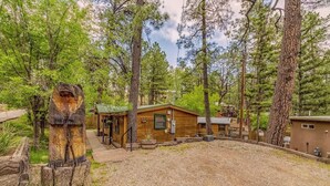 A rustic cabin surrounded by tall pine trees, with a gravel driveway and a wooden fence.