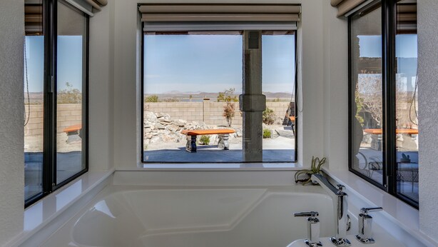 A bathroom with a large bathtub, a window with a view of a desert landscape, and a stone wall. - New Roadrunner heights (Joshua Tree)
