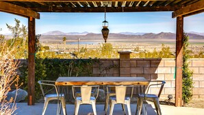 An outdoor patio with a wooden table and metal chairs, overlooking a desert landscape with mountains in the distance. - New Roadrunner heights (Joshua Tree)