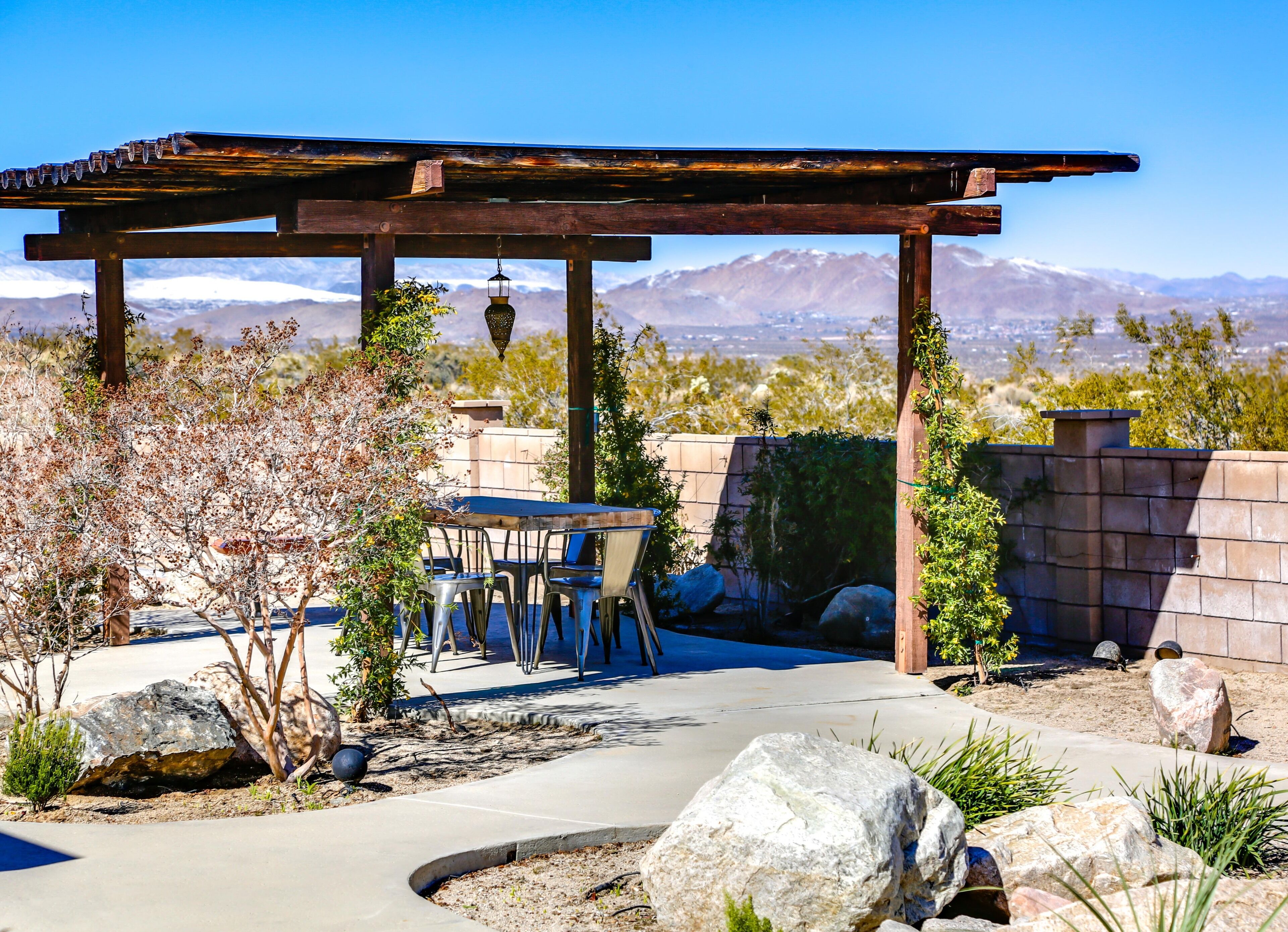 A covered patio with a table and chairs, surrounded by rocks and desert vegetation, overlooking a mountainous landscape.