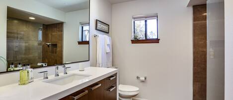 A modern bathroom with a large mirror, two sinks, a toilet, and a shower area.