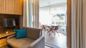Un salón moderno con un sofá, una mesa de centro y una televisión montada en una pared de madera. Se ve un balcón con una mesa y sillas a través de las puertas de cristal.