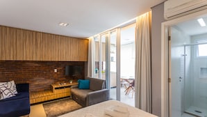 Una habitación de hotel moderna con pared de ladrillo decorativa, un sofá, un escritorio con televisor, una cama con toallas y una puerta corrediza de vidrio que da a un balcón.