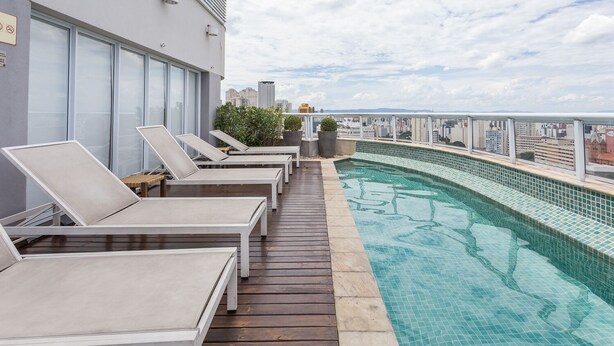 Un área de piscina en la azotea con sillones, piscina con baldosas y vistas a la ciudad.
