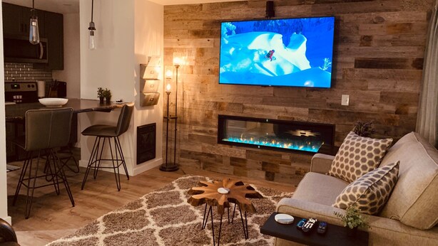 Ein modernes Wohnzimmer mit einem flachbildfernseher an einer Holzwand, einem Kamin, einer Couch mit gemusterten Kissen und einem Couchtisch mit Fernbedienungen.