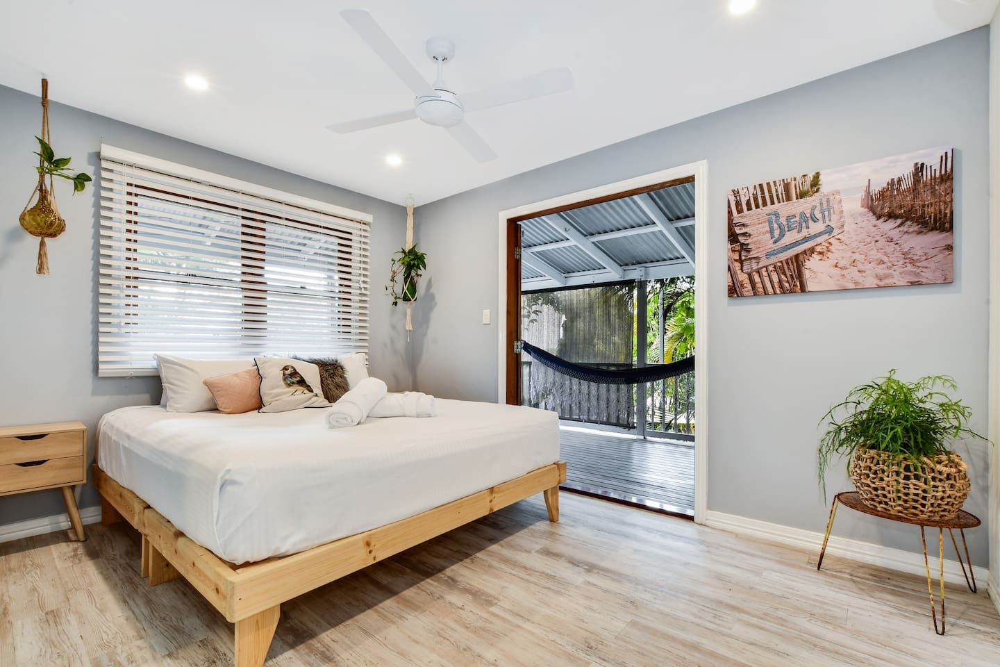A bedroom with a bed, a ceiling fan, a window with blinds, a hammock, and a beach-themed wall art.