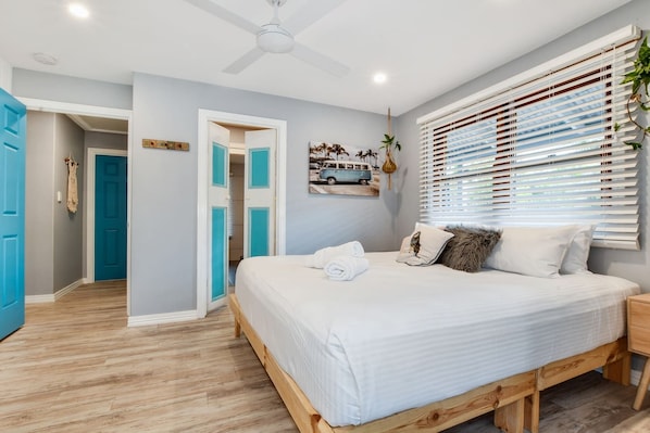 Un dormitorio con cama, ventilador de techo, ventana con persianas y puerta que da a otra habitación.
