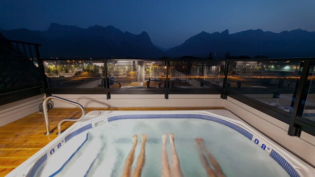 A hot tub on a balcony with a view of mountains and a cityscape at night. - Basecamp Resorts Canmore I One Bedroom Apartment, Dog Friendly (Canmore)
