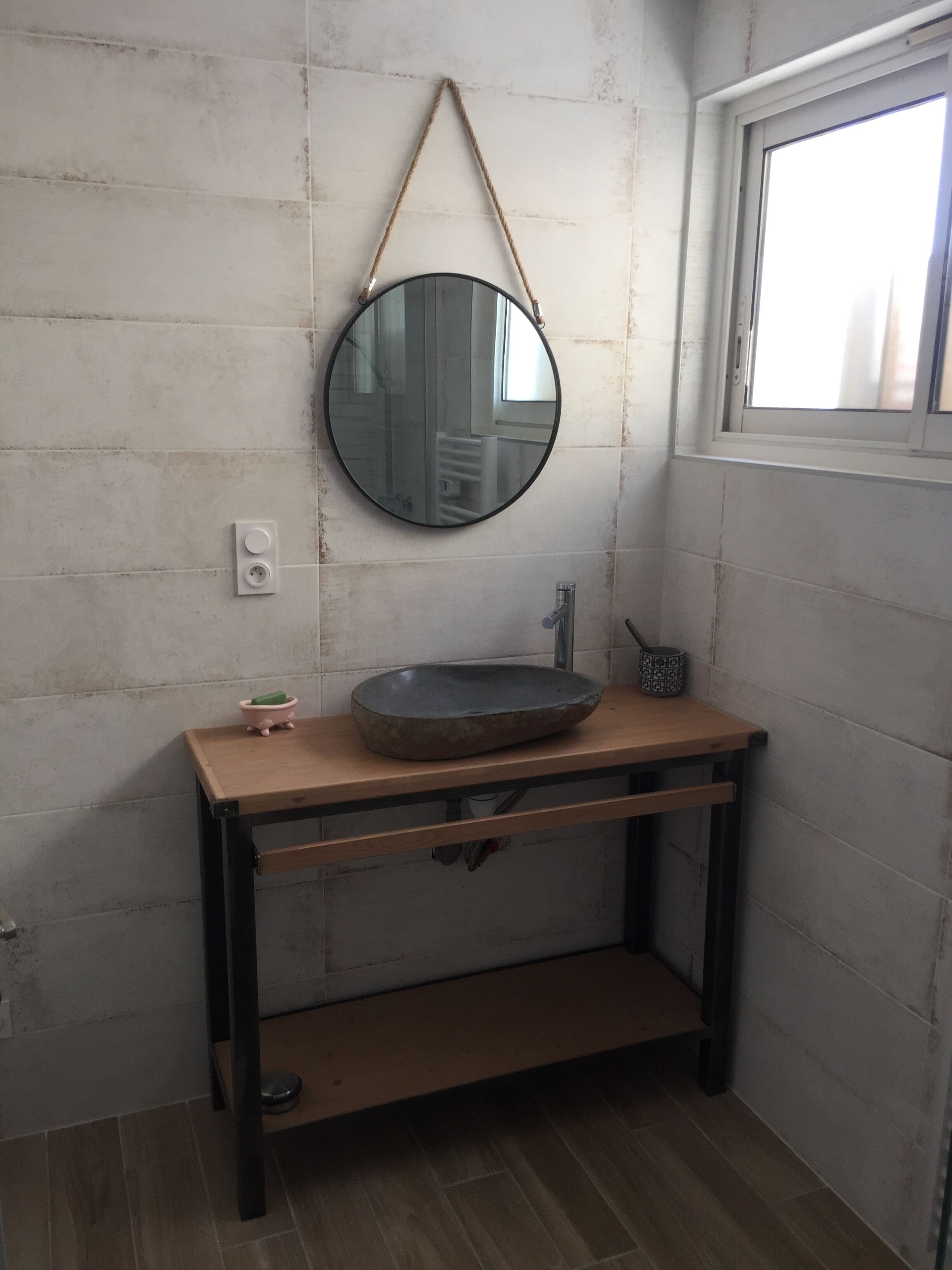 A bathroom with a round mirror, a wooden sink, and a window.