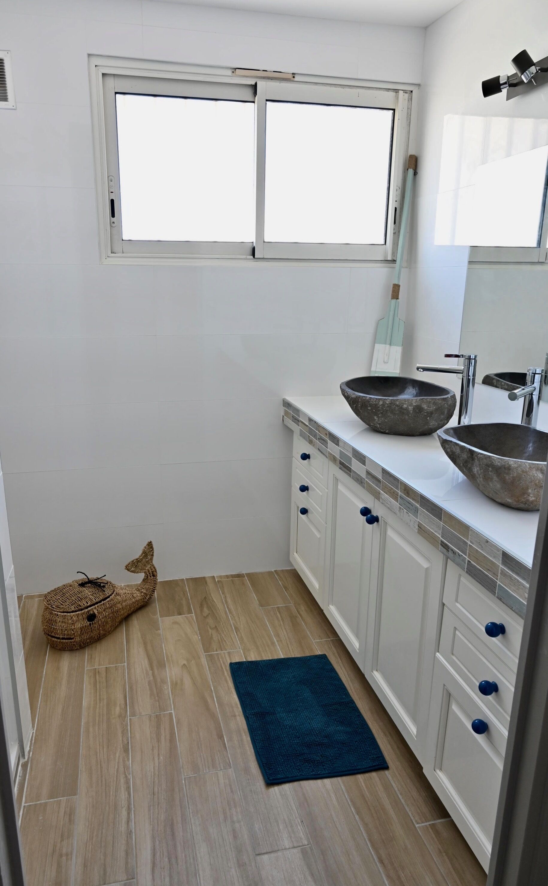 Une salle de bain moderne avec des meubles blancs, deux grandes fenêtres, un lavabo en pierre et un tapis bleu.