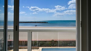 Uitzicht vanaf een balkon op een strand met een rotsachtige zeewering, een zwembad en een heldere lucht.