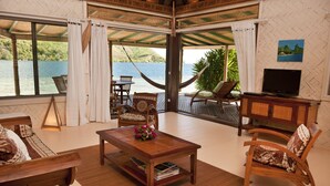 A room with a large window overlooking a body of water, a hammock, and a television on a wooden cabinet. - Beach Villa Cook - Robinson's Cove (Papetoai)