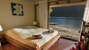 A bedroom with a bed, a nightstand, a window with a view of a beach and water. - Beach Villa Cook - Robinson's Cove (Papetoai)