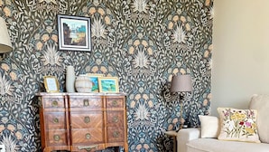 A living room with a patterned wallpaper, a wooden cabinet with decorative items, a beige sofa, and two glass-top coffee tables. - Avignon Lovely Flat (Villeneuve-lès-Avignon)