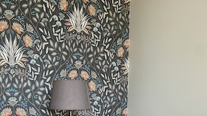 A room with a patterned wallpaper, a lamp on a side table, and a sofa with a floral cushion. - Avignon Lovely Flat (Villeneuve-lès-Avignon)
