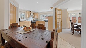 A spacious dining area with a wooden table and chairs, a kitchen in the background, and a view of the outside through a large window. - Taigh d'Luxe - Classic elegance meets modern chic in stunning spacious villa (Inverness)
