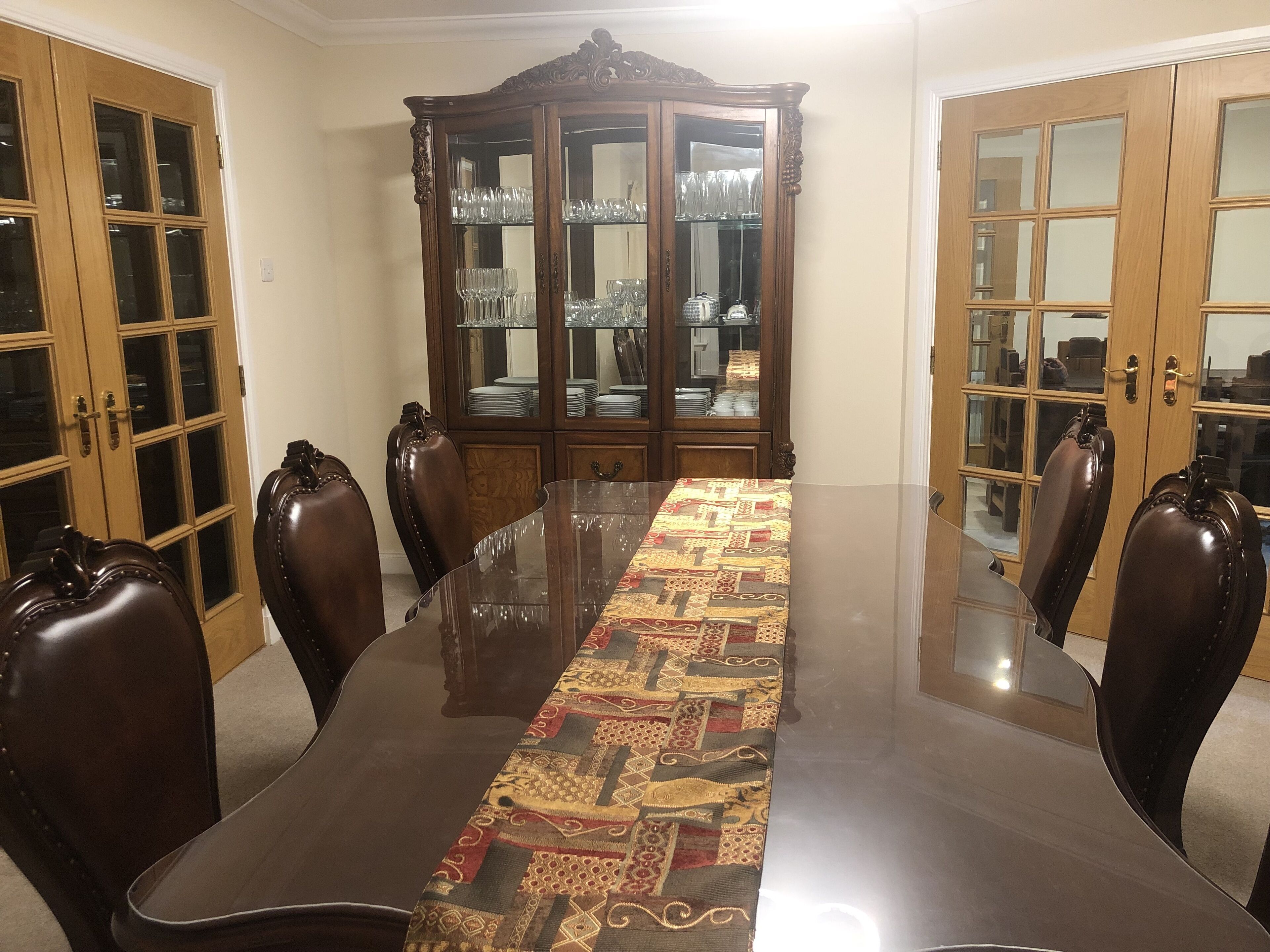 Une salle à manger avec une table à plateau en verre, un chemin de table à motifs, des chaises en cuir foncé et un buffet en bois avec des portes en verre.