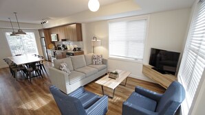 A modern living room with a sofa, two armchairs, a coffee table, and a TV. There is a dining area with a table and chairs, and a kitchen in the background. - Basecamp Resorts Canmore I Two Bedroom Apartment, Dog Friendly (Canmore)