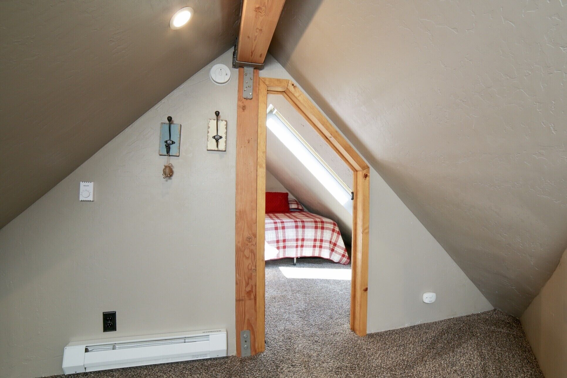 A small attic bedroom with a sloped ceiling, a wooden beam structure, a bed with a plaid bedspread, and a wall-mounted heater.