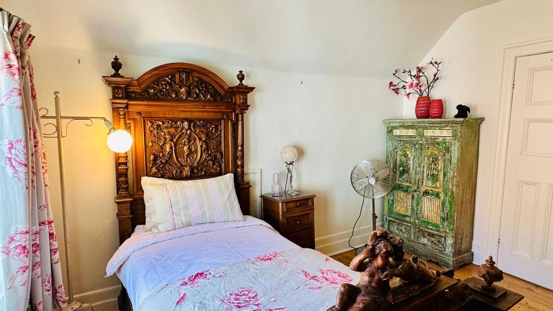 A bedroom with a wooden bed, a green cabinet, a nightstand with a lamp, and a fan.