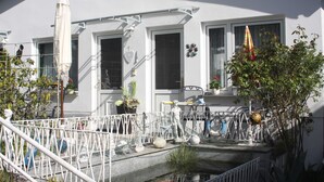 Um edifício branco com várias janelas, área de estar coberta ao ar livre e diversas decorações externas, incluindo uma estátua, uma bicicleta e um pequeno jardim.