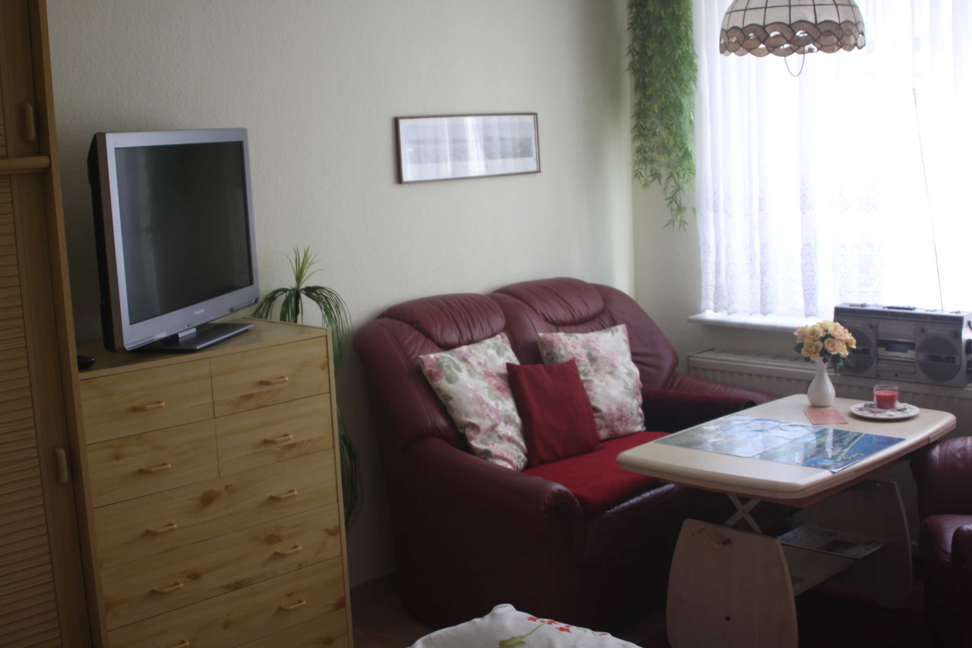 A living room with a television on a wooden cabinet, a red leather sofa with floral cushions, a small table with a vase and a drink, and a window with curtains.