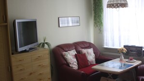 Uma sala de estar com televisão sobre um armário de madeira, um sofá de couro vermelho com almofadas florais, uma mesa pequena com um vaso e uma bebida, e uma janela com cortinas.