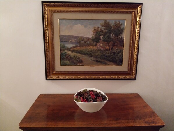 A framed landscape painting on a wall above a wooden table with a white bowl containing berries.