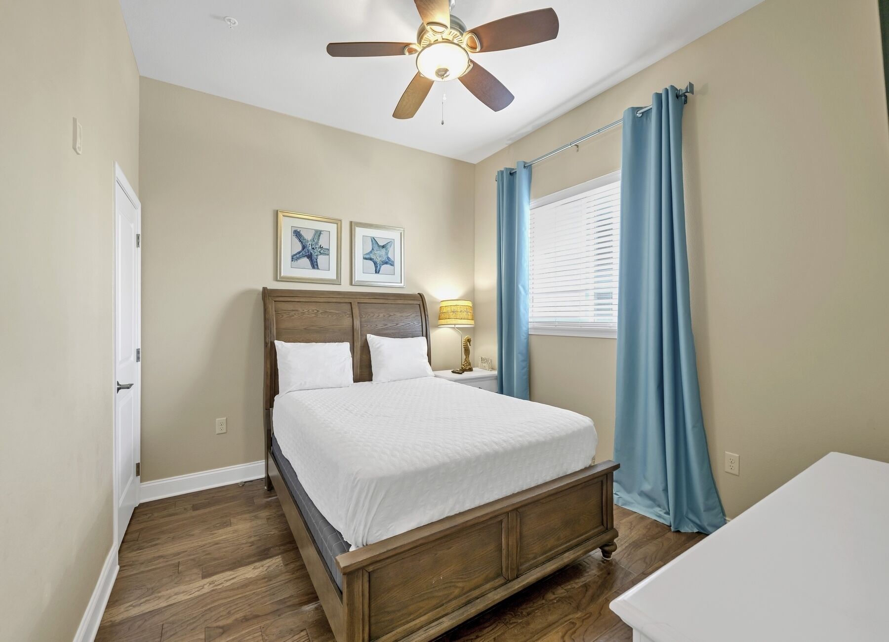 A bedroom with a wooden bed, a ceiling fan, a nightstand with a lamp, and two framed pictures on the wall.