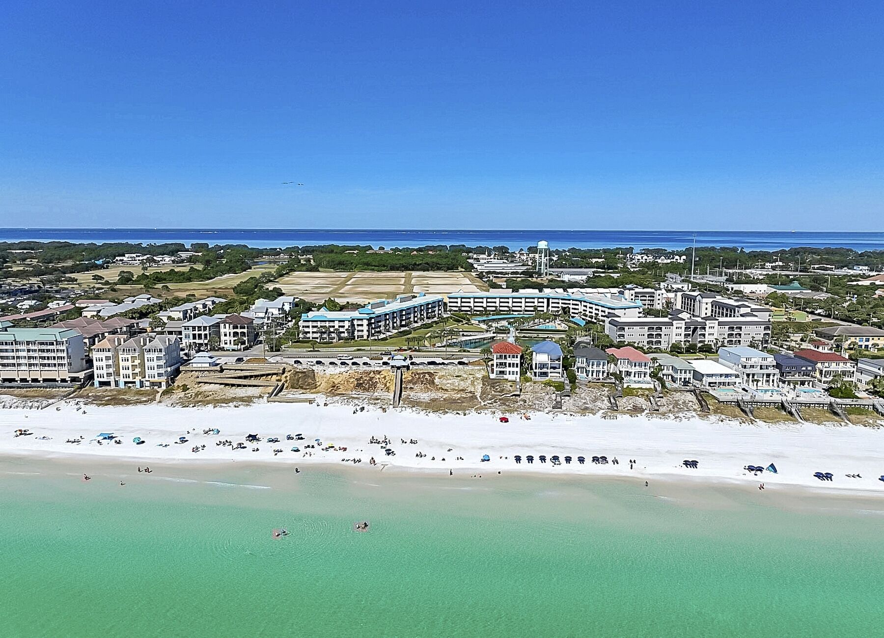 An aerial view of a coastal resort with multiple buildings, a beach, and a clear blue sky.