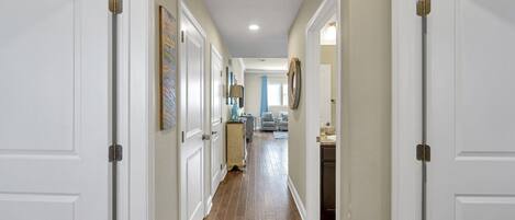 A hallway with white doors, wooden flooring, and a view into a living area with a sofa and a lamp.