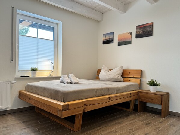 Une chambre à coucher avec un lit en bois, une fenêtre avec des stores, une table de chevet ornée d’une plante et deux tableaux accrochés au mur.