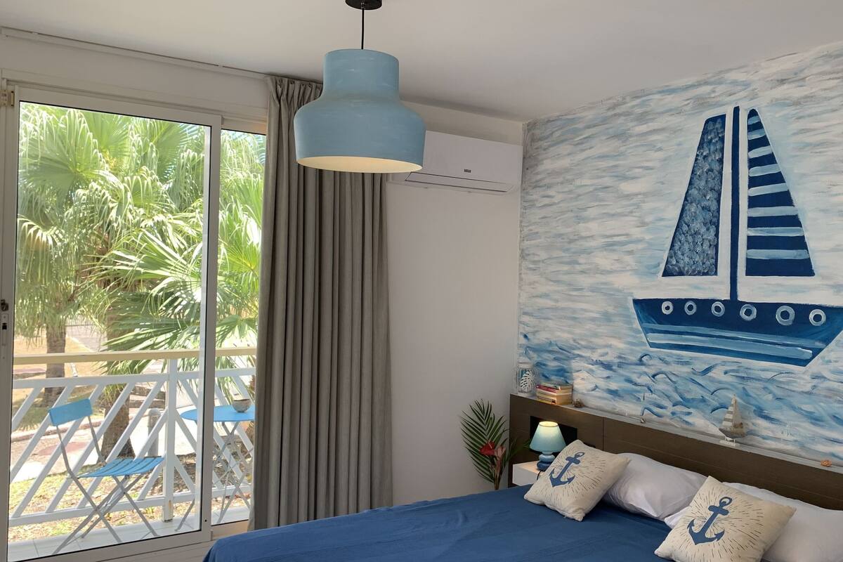 Un dormitorio con un ventanal grande, una cama con ropa de cama azul, una lámpara en el techo y un mural en la pared de un velero.
