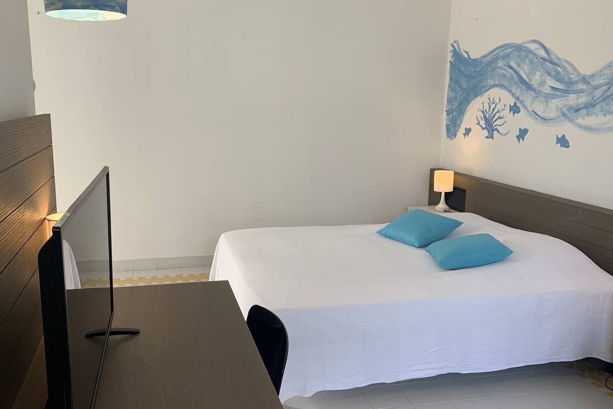Una habitación de hotel ordenada con una cama, mesita de noche, lámpara y un mural en la pared.
