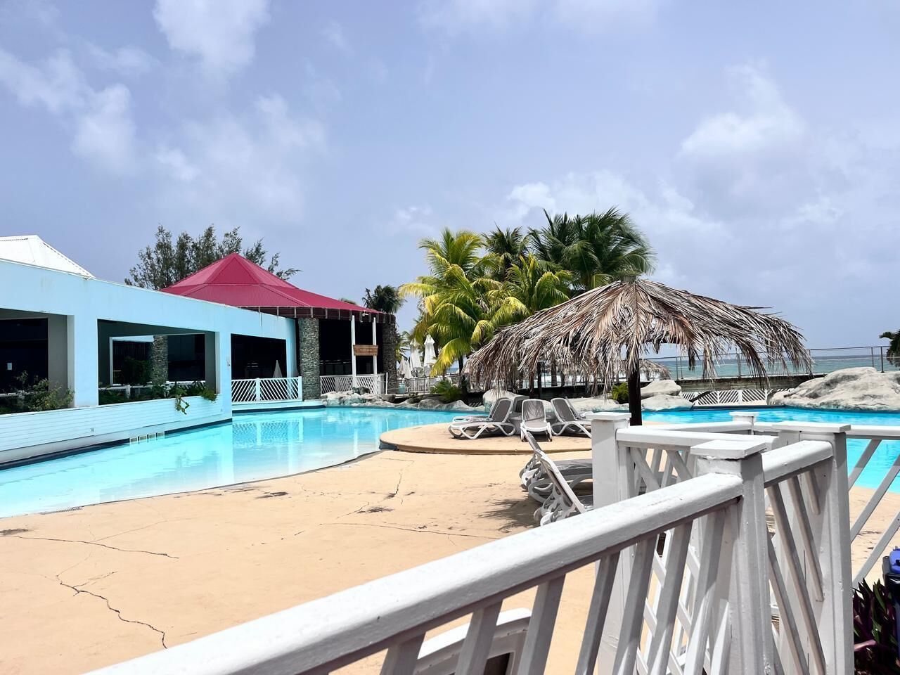 A pool area with lounge chairs, a thatched umbrella, and a building with a red roof.